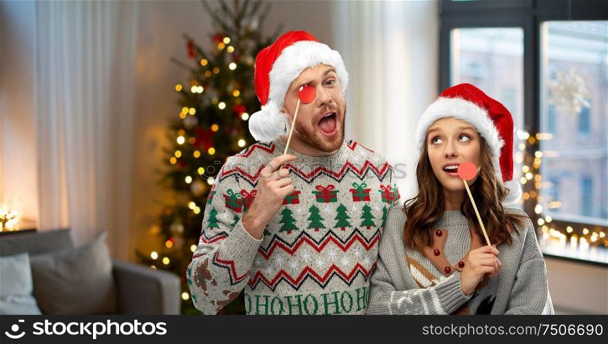 christmas, photo booth and holidays concept - happy couple in ugly sweaters posing with party props over home room background. couple with christmas party props in ugly sweaters