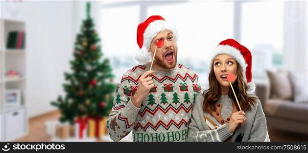 christmas, photo booth and holidays concept - happy couple in ugly sweaters posing with party props over home room background. couple with christmas party props in ugly sweaters