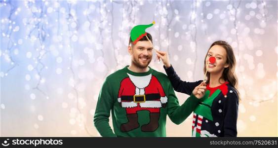 christmas, photo booth and holidays concept - happy couple in ugly sweaters posing with party props over festive lights background. couple with christmas party props in ugly sweaters