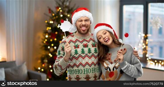 christmas, photo booth and holidays concept - happy couple in ugly sweaters posing with party props over home background. couple with christmas party props in ugly sweaters