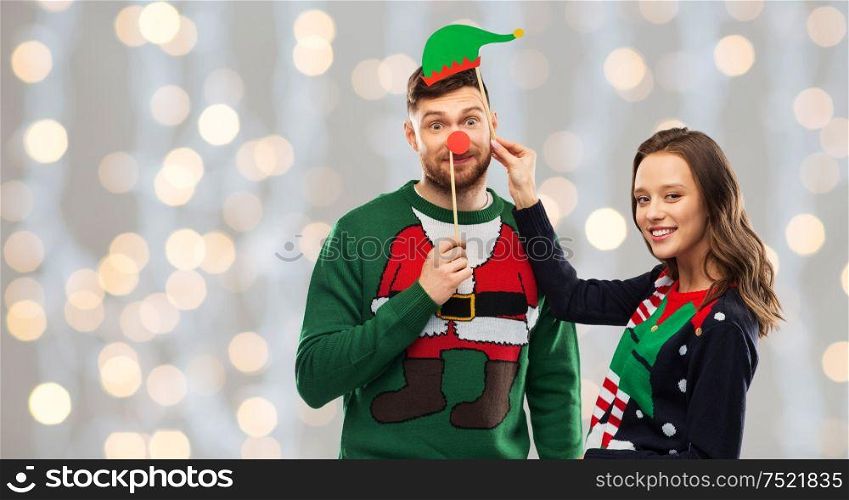christmas, photo booth and holidays concept - happy couple in ugly sweaters posing with party props over festive lights background. couple with christmas party props in ugly sweaters