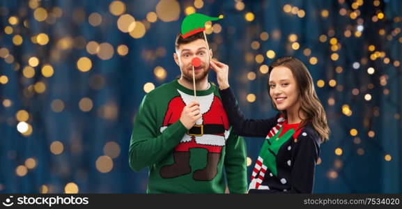 christmas, photo booth and holidays concept - happy couple in ugly sweaters posing with party props over festive lights on dark night background. couple with christmas party props in ugly sweaters
