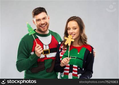 christmas, photo booth and holidays concept - happy couple in ugly sweaters posing with party props. couple with christmas party props in ugly sweaters