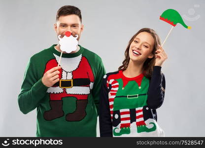 christmas, photo booth and holidays concept - happy couple in ugly sweaters posing with party props. couple with christmas party props in ugly sweaters