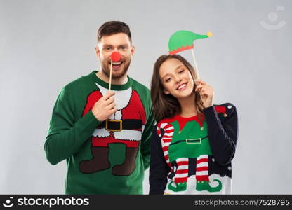 christmas, photo booth and holidays concept - happy couple in ugly sweaters posing with party props. couple with christmas party props in ugly sweaters