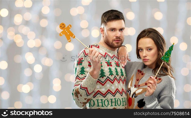 christmas, photo booth and holidays concept - sad couple in ugly sweaters posing with party props over festive lights background. couple with christmas party props in ugly sweaters