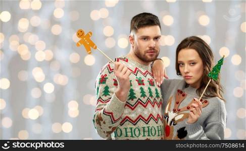christmas, photo booth and holidays concept - sad couple in ugly sweaters posing with party props over festive lights background. couple with christmas party props in ugly sweaters