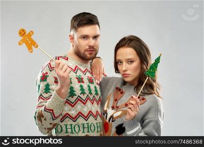 christmas, photo booth and holidays concept - sad couple in ugly sweaters posing with party props. couple with christmas party props in ugly sweaters