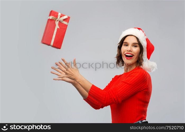 christmas, presents and holidays concept - happy smiling young woman in santa helper hat catching red gift box over grey background. happy young woman in santa hat catching gift box