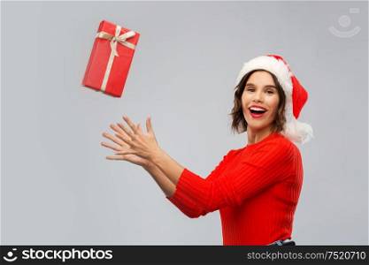 christmas, presents and holidays concept - happy smiling young woman in santa helper hat catching red gift box over grey background. happy young woman in santa hat catching gift box