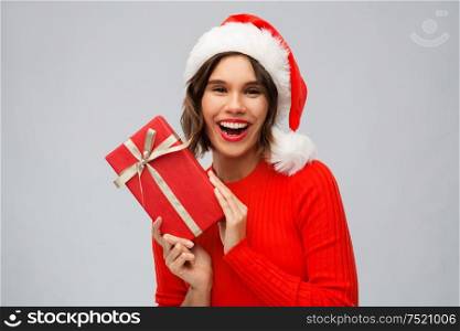 christmas, presents and holidays concept - happy smiling young woman in santa helper hat with red gift box over grey background. happy young woman in santa hat with red gift box