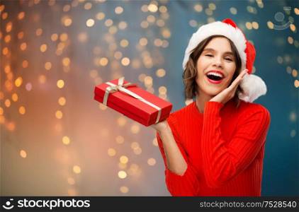christmas, presents and holidays concept - happy smiling young woman in santa helper hat with red gift box over festive lights background. happy young woman in santa hat with christmas box