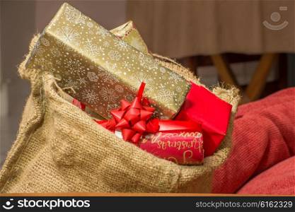 Christmas presents stacked to the brim inside a brown hessian bag resting near some red pillows inside a home.