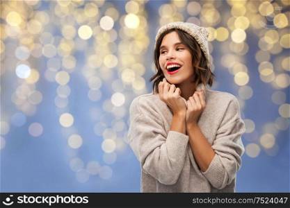 christmas, season and holidays concept - happy smiling young woman in knitted winter hat and sweater over festive lights on blue background. young woman in winter hat and sweater on christmas