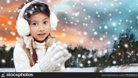 christmas, season and people concept - happy little girl wearing earmuffs over winter forest background. happy little girl in earmuffs over winter forest
