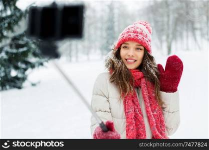 christmas, season and people concept - happy smiling teenage girl or woman taking picture by selfie stick and waving hand in winter park. young woman taking selfie by monopod in winter
