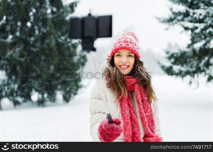christmas, season and people concept - happy smiling teenage girl or woman taking picture by selfie stick in winter park. young woman taking selfie by monopod in winter
