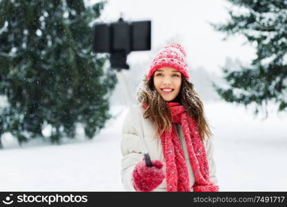 christmas, season and people concept - happy smiling teenage girl or woman taking picture by selfie stick in winter park. young woman taking selfie by monopod in winter