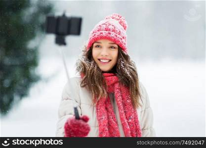 christmas, season and people concept - happy smiling teenage girl or woman taking picture by selfie stick in winter park. young woman taking selfie by monopod in winter