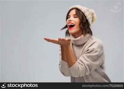 christmas, season and people concept - happy smiling young woman in knitted winter hat and sweater sending air kiss over grey background. young woman in knitted winter hat sending air kiss