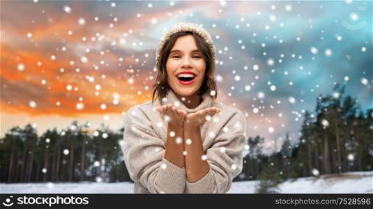 christmas, season and people concept - happy smiling young woman in knitted hat and sweater sending air kiss over winter forest and snow background. woman in hat sending air kiss over winter forest