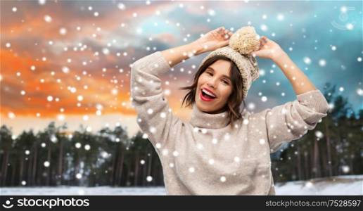 christmas, season and people concept - happy smiling young woman in knitted hat and sweater over winter forest and snow background. woman in hat and sweater over winter forest