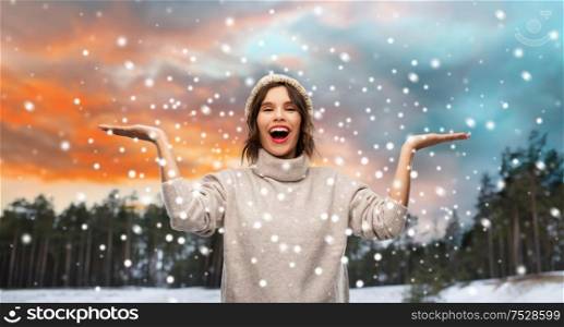 christmas, season and people concept - happy smiling young woman in knitted hat and sweater holding something on empty hand palm over winter forest and snow background. woman in hat and sweater over winter forest