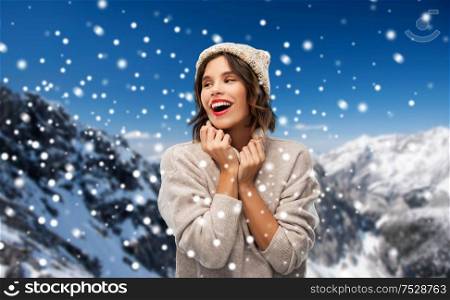 christmas, season and people concept - happy smiling young woman in knitted winter hat and sweater over snow and alps mountains background. young woman in knitted winter hat in mountains