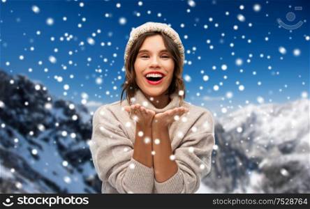 christmas, season and people concept - happy smiling young woman in knitted winter hat and sweater sending air kiss over snow and alps mountains background. young woman in knitted winter hat in mountains