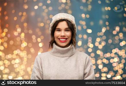 christmas, season and people concept - happy smiling young woman in knitted winter hat and sweater over festive lights background. young woman in knitted winter hat and sweater