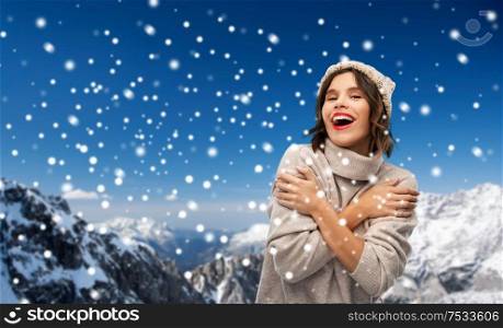 christmas, season and people concept - happy smiling young woman in knitted winter hat and sweater over snow and alps mountains background. young woman in knitted winter hat and sweater