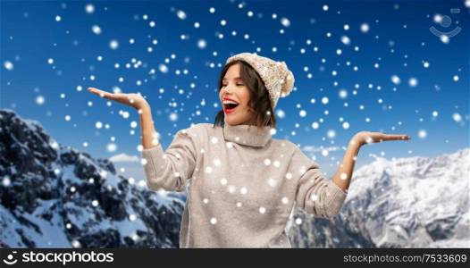 christmas, season and people concept - happy smiling young woman in knitted winter hat and sweater holding something on empty hand palm over snow and alps mountains background. young woman in winter hat holding something