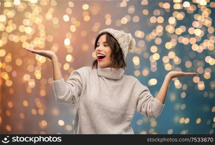 christmas, season and people concept - happy smiling young woman in knitted winter hat and sweater holding something on empty hand palm over festive lights background. young woman in winter hat holding something