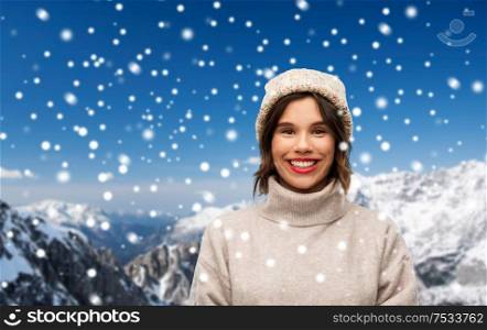 christmas, season and people concept - happy smiling young woman in knitted winter hat and sweater over snow and alps mountains background. young woman in knitted winter hat and sweater