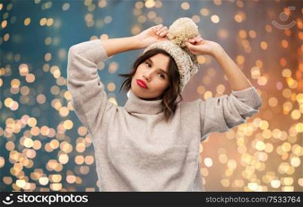christmas, season and people concept - happy smiling young woman in knitted winter hat and sweater over festive lights background. young woman in knitted winter hat and sweater
