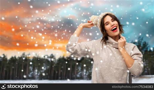 christmas, season and people concept - happy smiling young woman in knitted hat and sweater over winter forest and snow background. woman in hat and sweater over winter forest