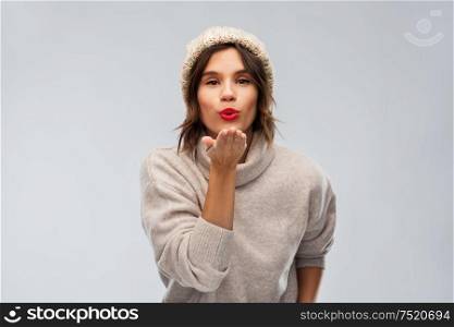 christmas, season and people concept - happy young woman in knitted winter hat and sweater sending air kiss over grey background. young woman in knitted winter hat sending air kiss