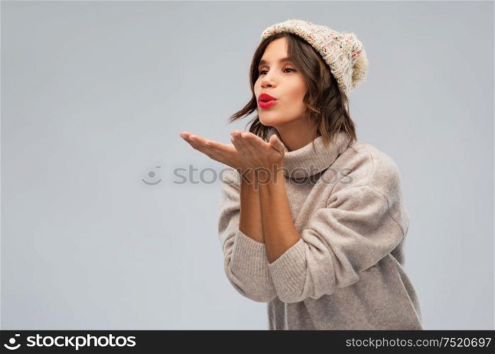 christmas, season and people concept - happy young woman in knitted winter hat and sweater sending air kiss over grey background. young woman in knitted winter hat sending air kiss