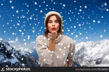 christmas, season and people concept - happy young woman in knitted winter hat and sweater sending air kiss over snow and alps mountains background. young woman in knitted winter hat sending air kiss