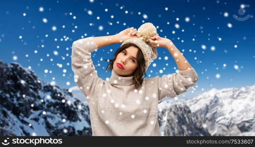 christmas, season and people concept - happy young woman in knitted winter hat and sweater over snow and alps mountains background. young woman in knitted winter hat and sweater