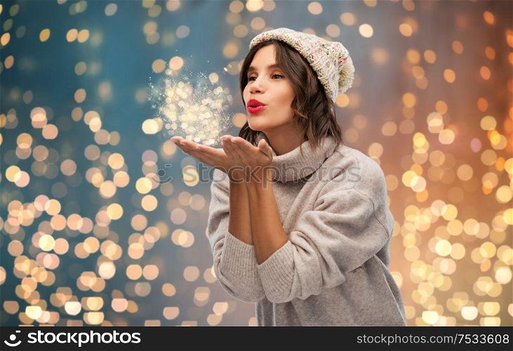 christmas, season and people concept - happy young woman in knitted winter hat and sweater sending air kiss over festive lights background. young woman in knitted winter hat sending air kiss