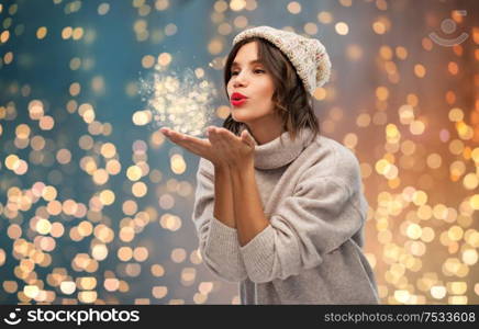 christmas, season and people concept - happy young woman in knitted winter hat and sweater sending air kiss over festive lights background. young woman in knitted winter hat sending air kiss