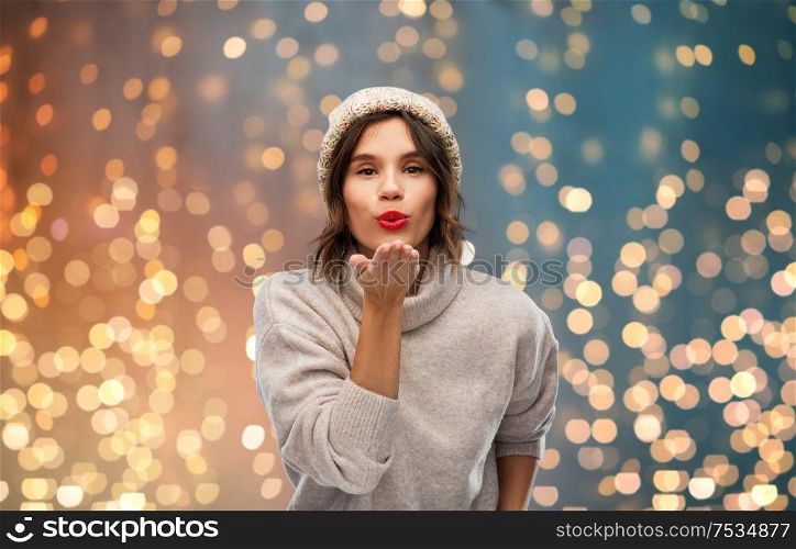 christmas, season and people concept - happy young woman in knitted winter hat and sweater sending air kiss over festive lights background. young woman in knitted winter hat sending air kiss