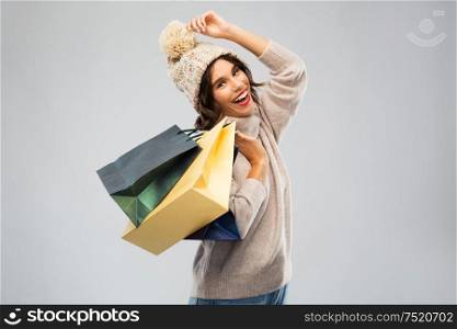 christmas, seasonal sale and consumerism concept - happy smiling young woman in knitted winter hat and sweater with shopping bags over grey background. young woman in winter hat with shopping bags
