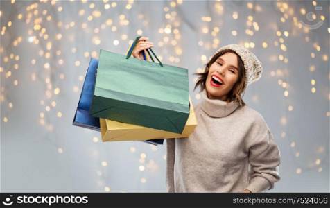 christmas, seasonal sale and consumerism concept - happy smiling young woman in knitted winter hat and sweater with shopping bags over festive lights on grey background. young woman in hat with shopping bags on christmas
