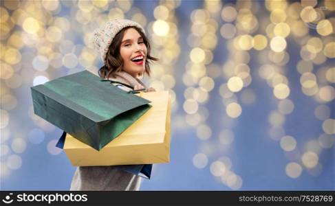christmas, seasonal sale and consumerism concept - happy smiling young woman in knitted winter hat and sweater with shopping bags over festive lights on blue background. woman in hat with shopping bags on christmas