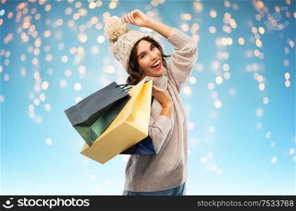 christmas, seasonal sale and consumerism concept - happy smiling young woman in knitted winter hat and sweater with shopping bags over festive lights on blue background. young woman in winter hat with shopping bags