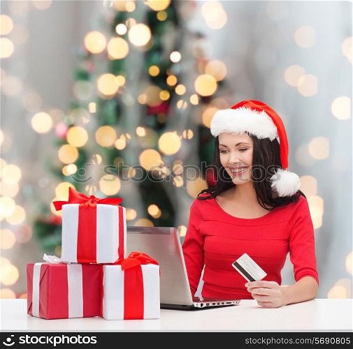 christmas, shopping and people concept - smiling woman in santa helper hat with gift boxes, laptop computer and credit card over tree lights background