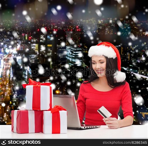 christmas, shopping and people concept - smiling woman in santa helper hat with gift boxes, laptop computer and credit card over snowy night city background