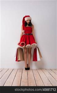 Christmas shopping. Pretty woman leaning on white wall with shopping bags, wearing red Santa Claus hat and dress